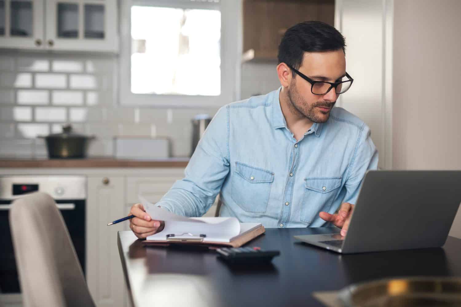 Young man typing on a laptop and doing paperwork from home.