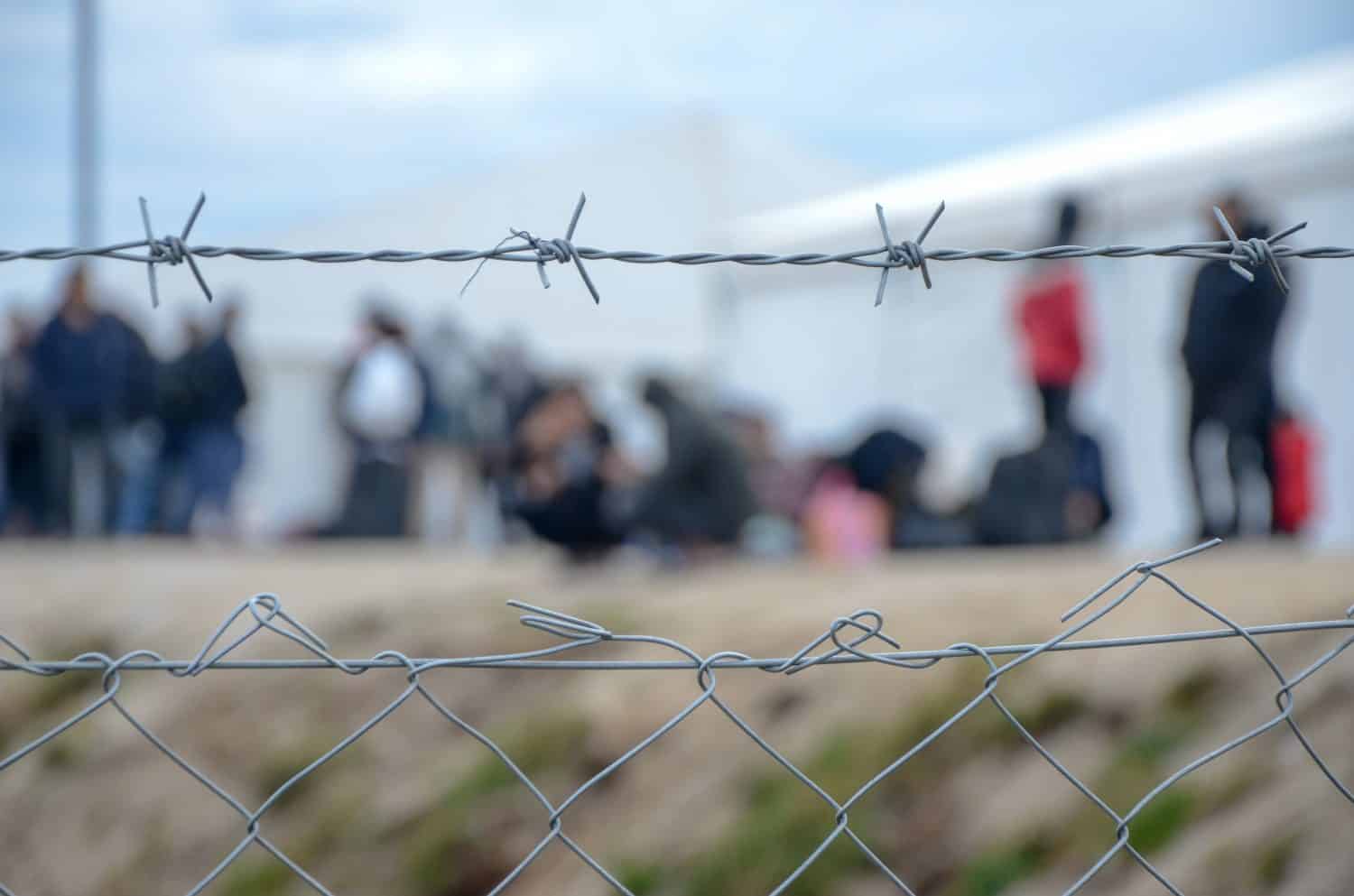 Barbed wire in refugee camp. Migrants behind chain link fence in camp. Group of people behind fence. Concept of prison, freedom, barrier, security and migration. Refugees on their way to EU.