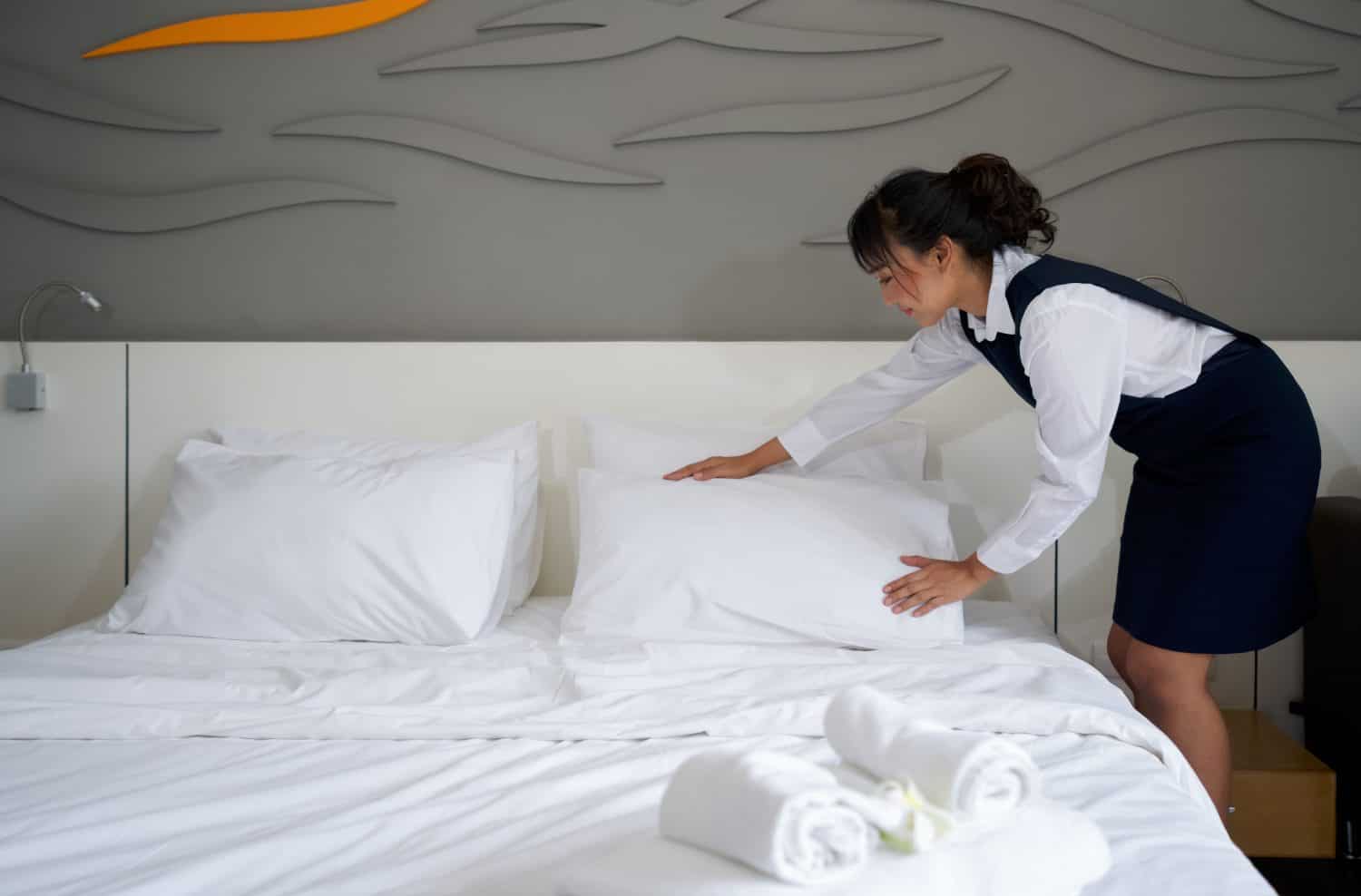 Young hotel maid making the bed. Hotel staff in blue uniform preparing room for guest.