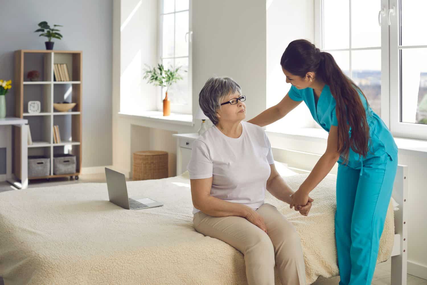 Trying to make patient&#039;s life happier. Female home care nurse supports and assists senior woman with all her daily needs. Caregiver holds old lady&#039;s hand, helps her stand up from bed and talks to her