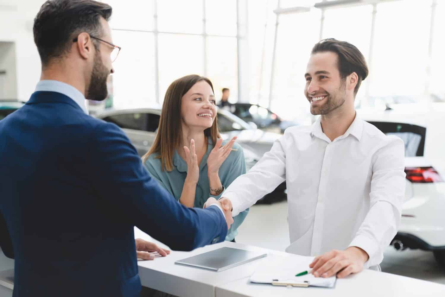 Great deal! Profitable deal handshake after buying purchasing new car. Male shop assistant congratulating young family couple with buying auto at dealer shop.