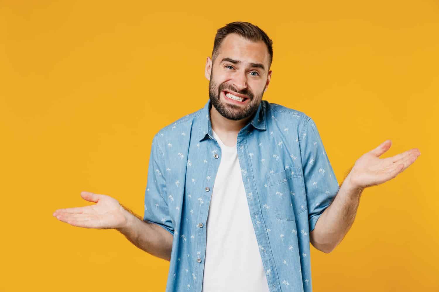 Young confused sad man 20s wearing blue shirt shrugging shoulders looking puzzled, have no idea, nothing to say, standing questioned and unaware isolated on plain yellow background studio portrait.