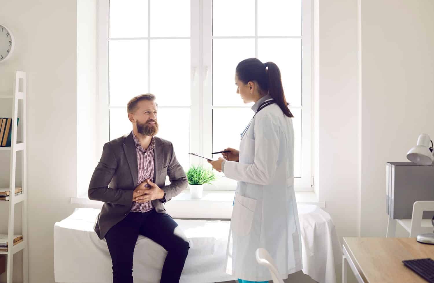 Patient with stomach ache visiting gastroenterologist. Unhappy young man suffering from gastritis stomachache having appointment, sitting on couch in modern exam room and asking doctor for help