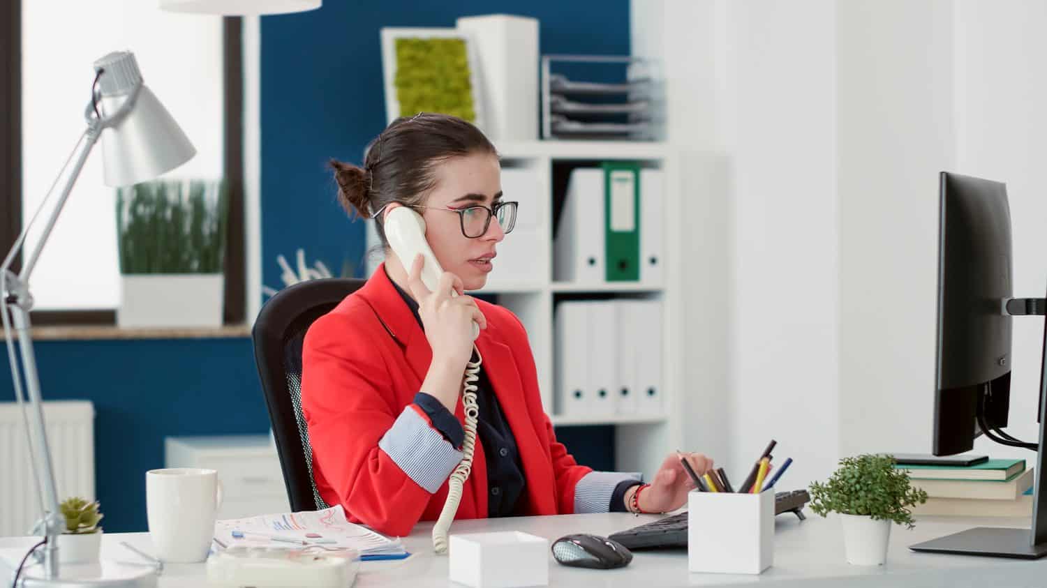 Company secretary having remote conversation on landline phone, working on financial development strategy with computer. Female employee answering office telephone call for startup work.