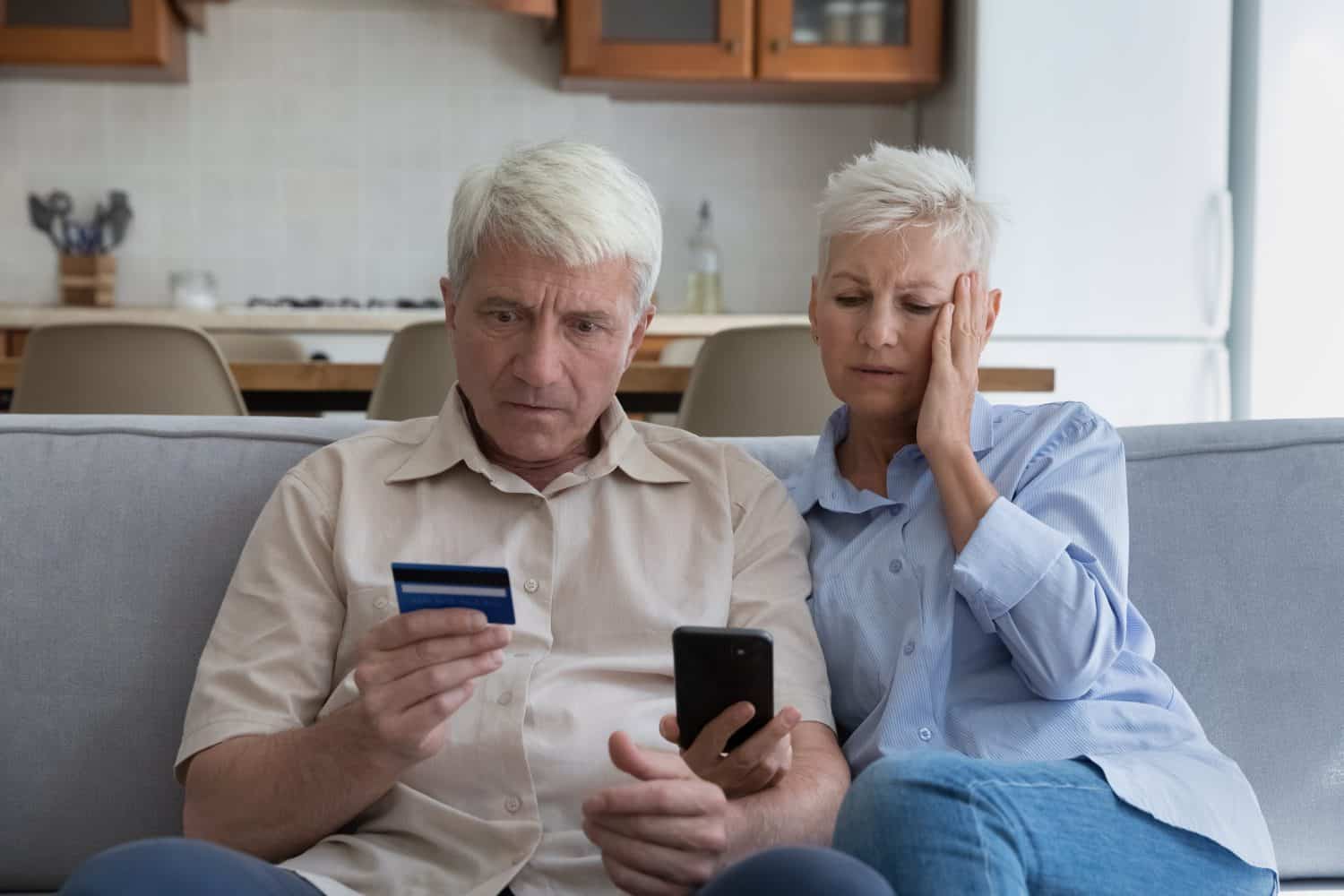 Shocked aged couple become victims of online fraud using credit card phone to pay for goods order service online on suspicious website. Frustrated older spouses overspending money at internet shopping