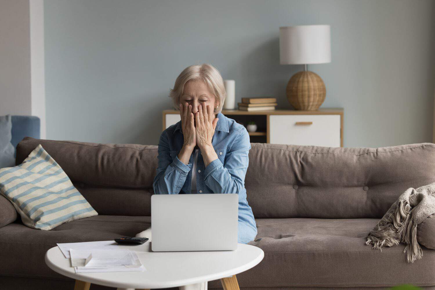 Shocked concerned senior accountant woman finding financial failure, mistake, bankruptcy, sitting at laptop, calculator, paper bills, covering face in panic attack, staring at screen