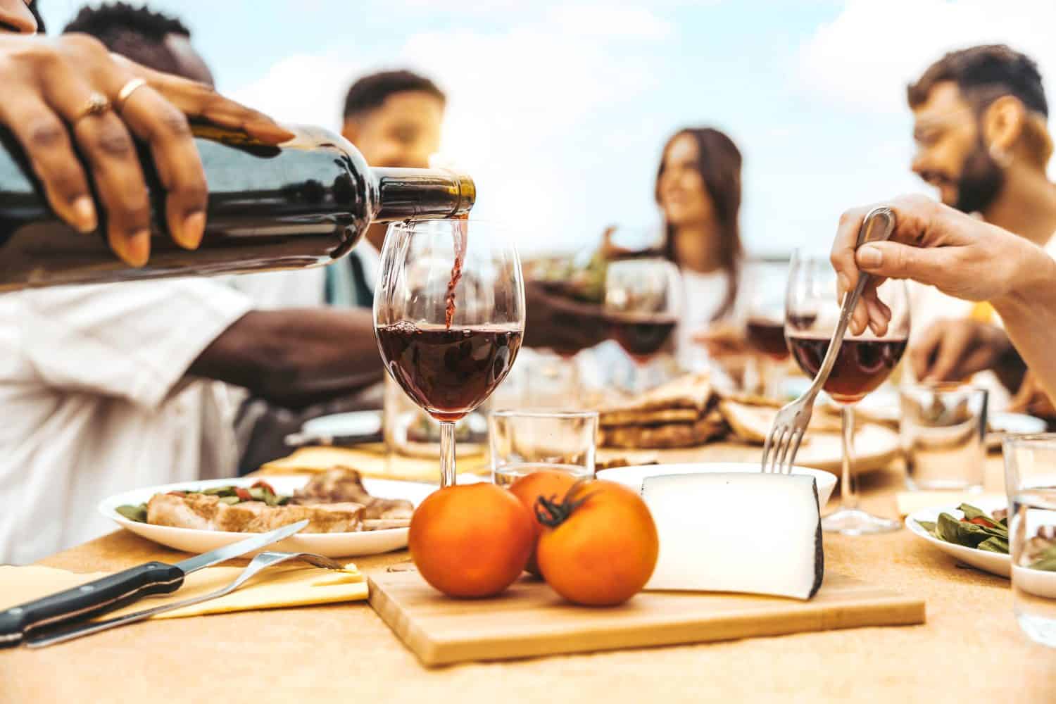 Happy friends drinking red wine sitting at winery restaurant balcony -Group of people having bbq dinner party outside - Food and beverage concept with guys and girls eating fresh meal at lunch break