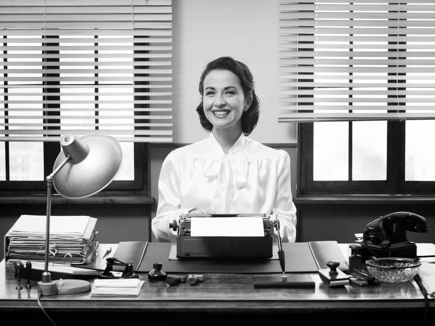 Cheerful vintage secretary working at office desk and smiling at camera