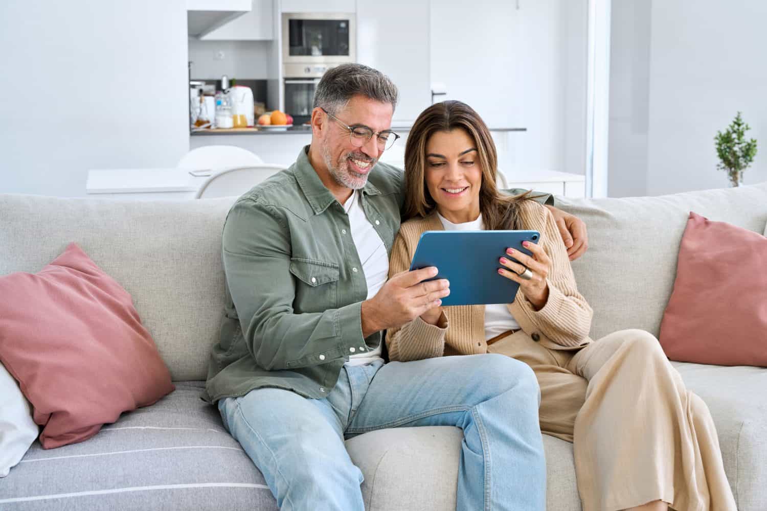 Happy middle aged couple using digital tablet relaxing on couch at home. Smiling mature man and woman holding tab computer browsing internet on pad technology device sitting on sofa in living room.