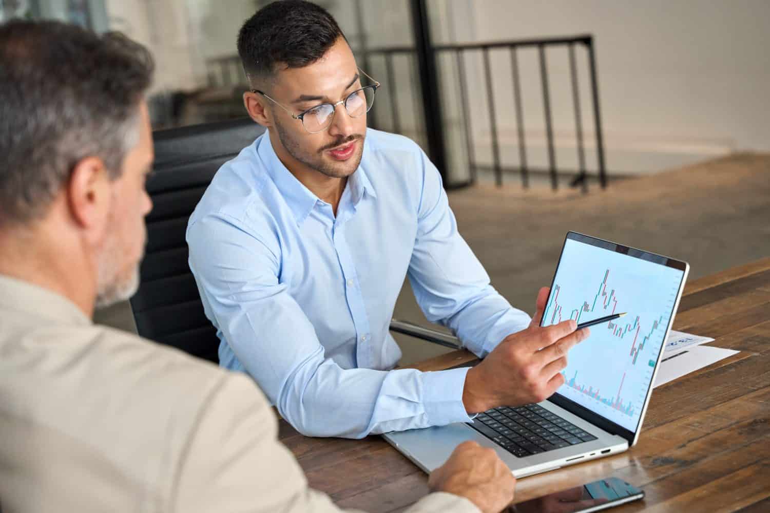 Financial advisor explaining invest stock market data consulting investor. Two busy business men analysts doing finance trading analysis pointing at exchange chart on laptop screen working in office.