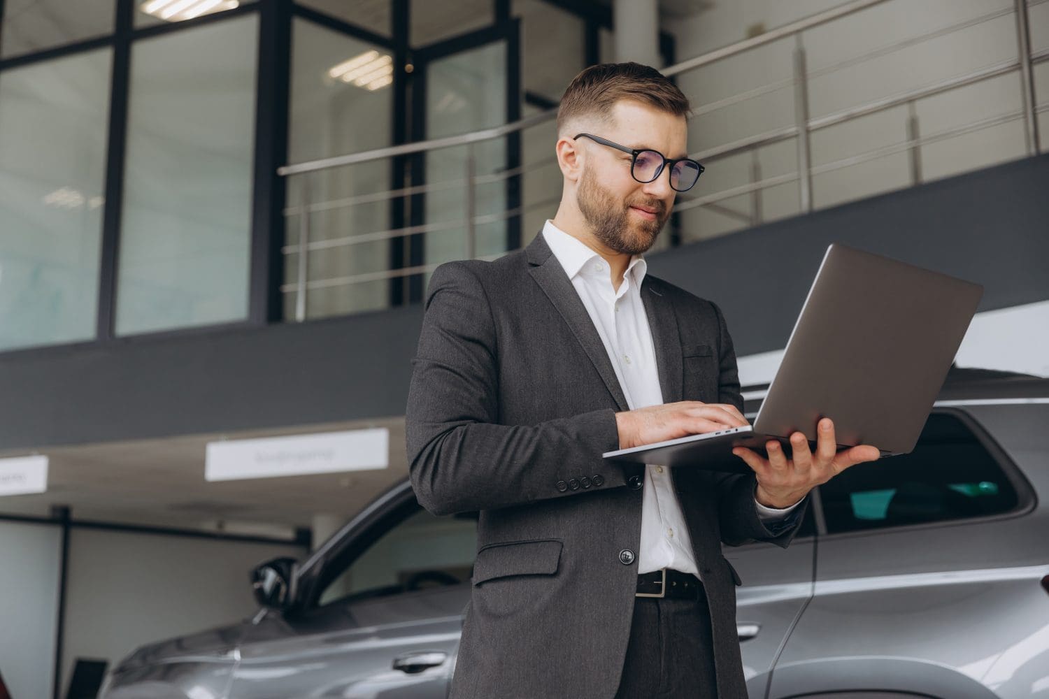 Modern bearded man in glasses and suit vehicle sales consultant using laptop inside car dealership