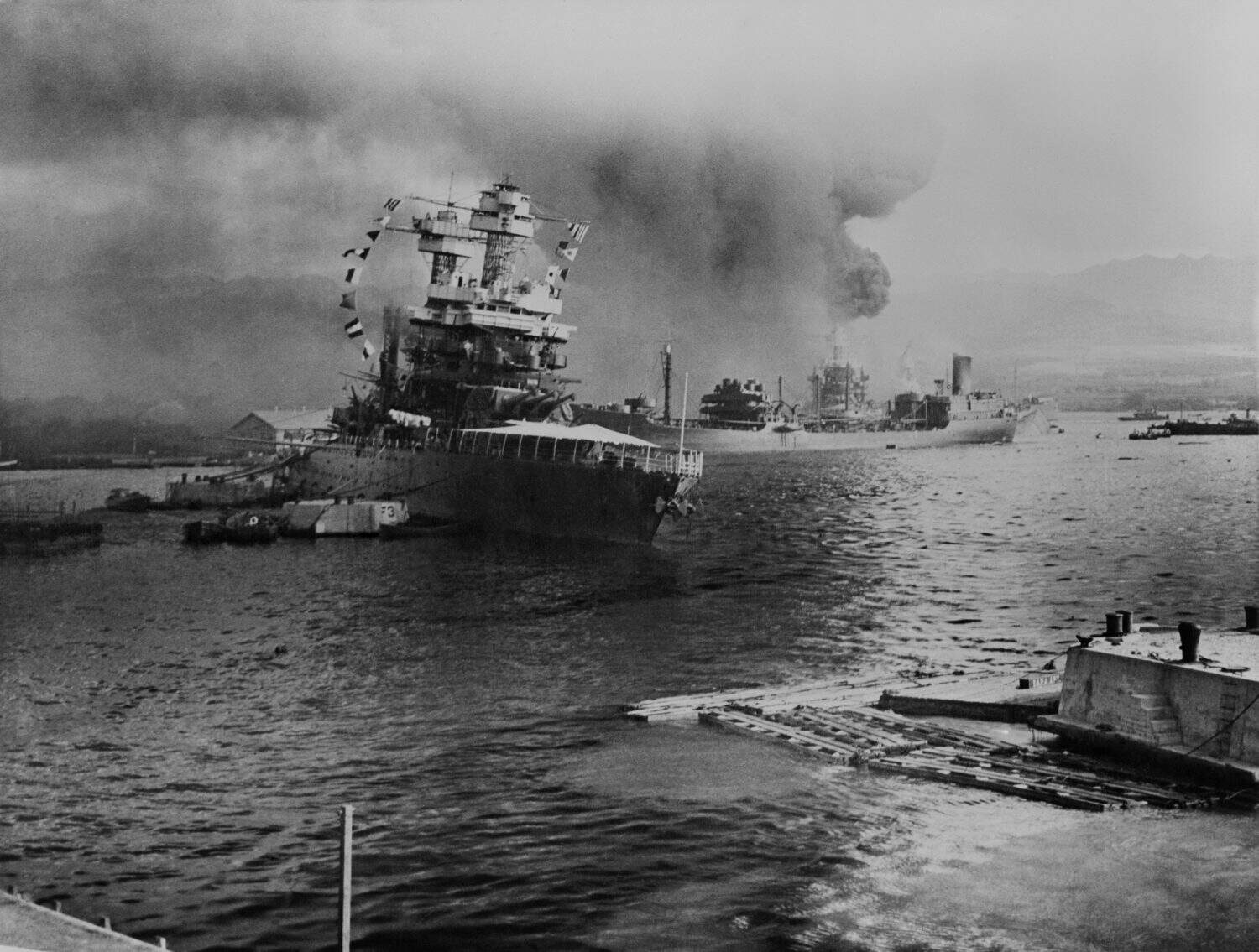 USS California lists after aerial blows taken during the Japanese attack on Pearl Harbor. In distance the USS Neosho, cautiously backs away from berth, avoiding capsized USS Oklahoma. Dec. 7, 1941.