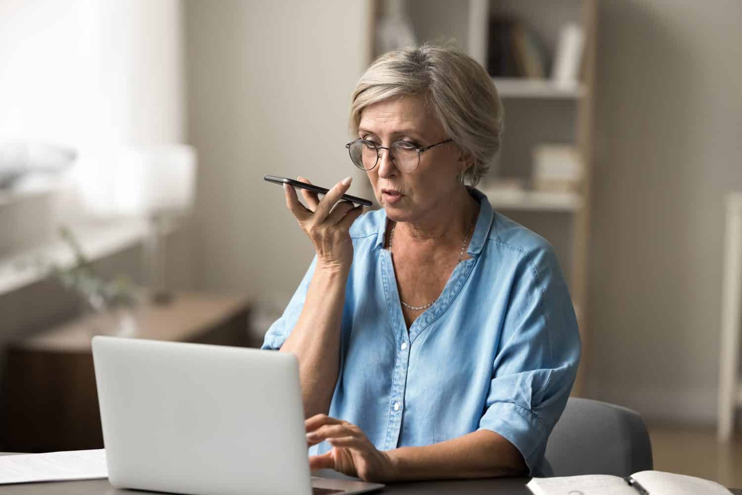 Mature woman using smartphone sit at desk with laptop, engaged in voice search or dictation on phone, input information, search data, managing work online, send audio message. Multitask, modern tech