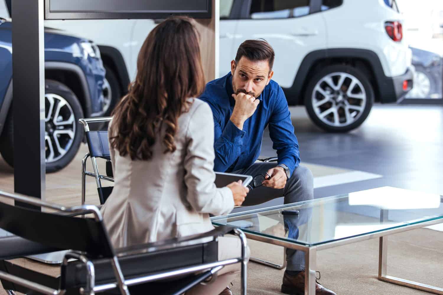 A man and woman engage in a focused business discussion inside a stylish car dealership. They are seated at a modern glass table, surrounded by new cars, suggesting decision-making and negotiation.