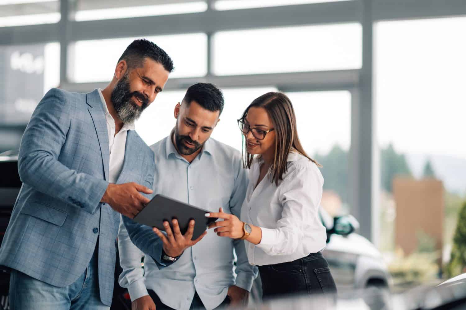 Dealer demonstrating digital information about a vehicle model to an attentive couple, utilizing a tablet, demonstrating the benefits and features, in an inviting showroom.