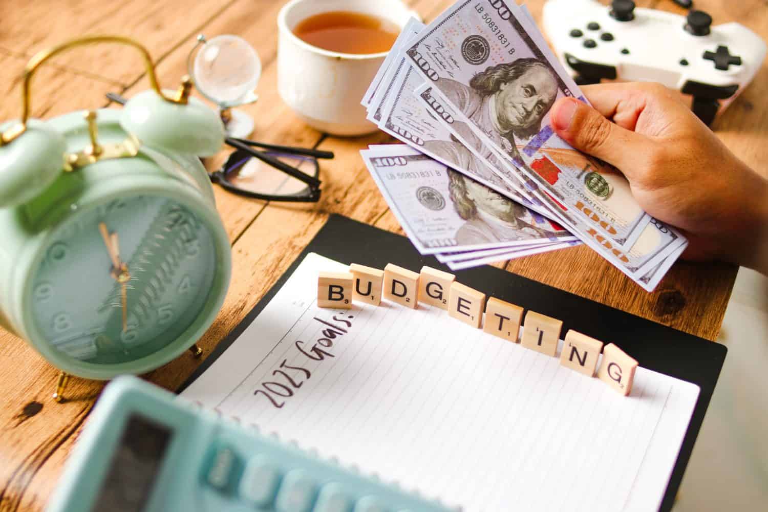 2025 Budgeting - A person planning their budget with money, alarm clock, tea, gamepad, and notebook, emphasizing the importance of financial planning and goal-setting for the future