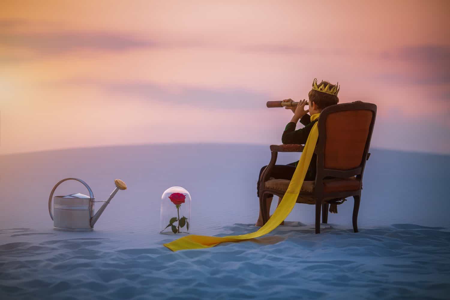 A boy dressed as the Little Prince sits on a chair in the desert, gazing through a telescope near a rose in a glass dome and a watering can. A whimsical and symbolic fantasy scene.