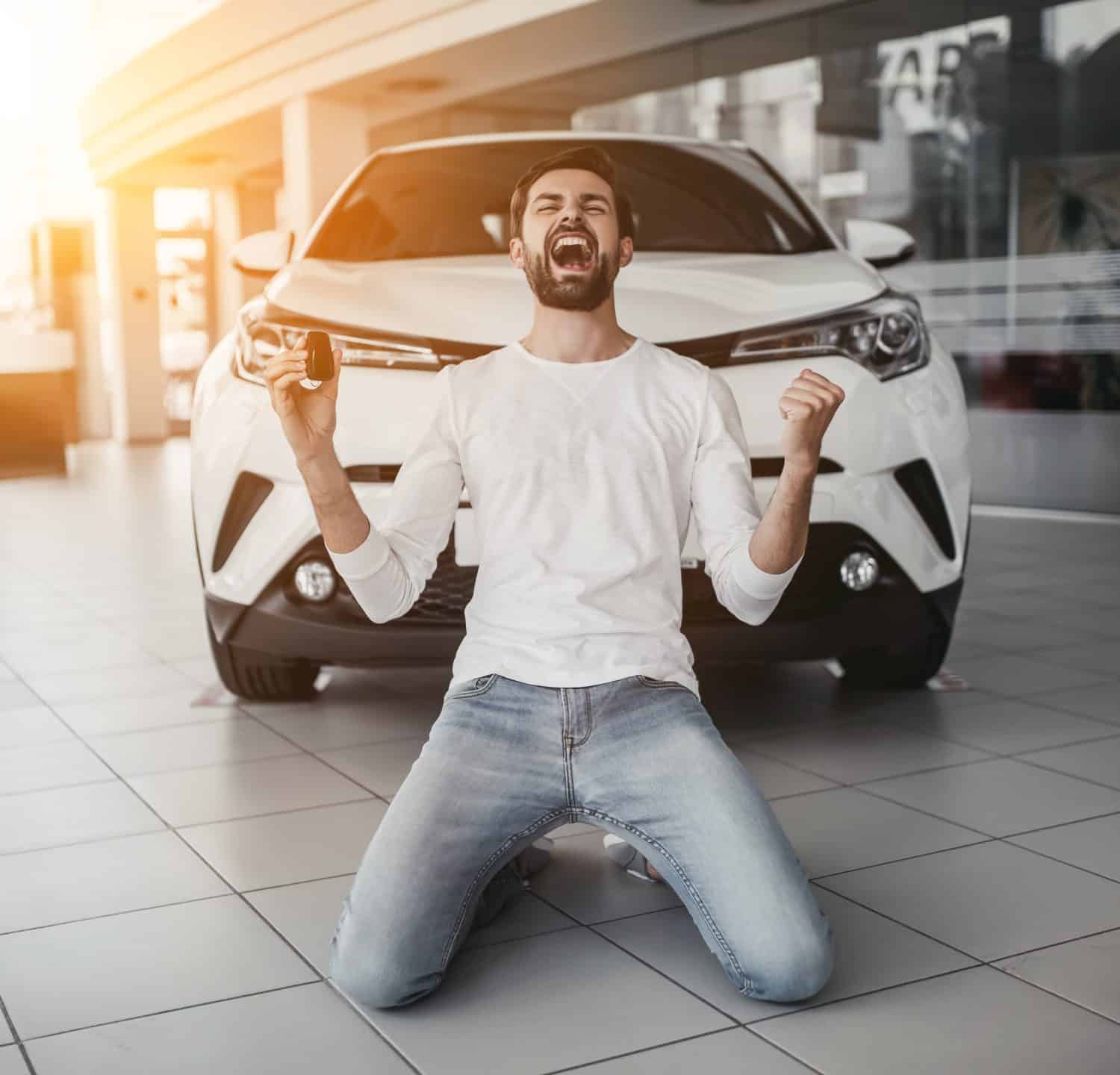 Yes, that&#039;s my new car! Customer in car dealership. Young man knelt down with keys in hand.