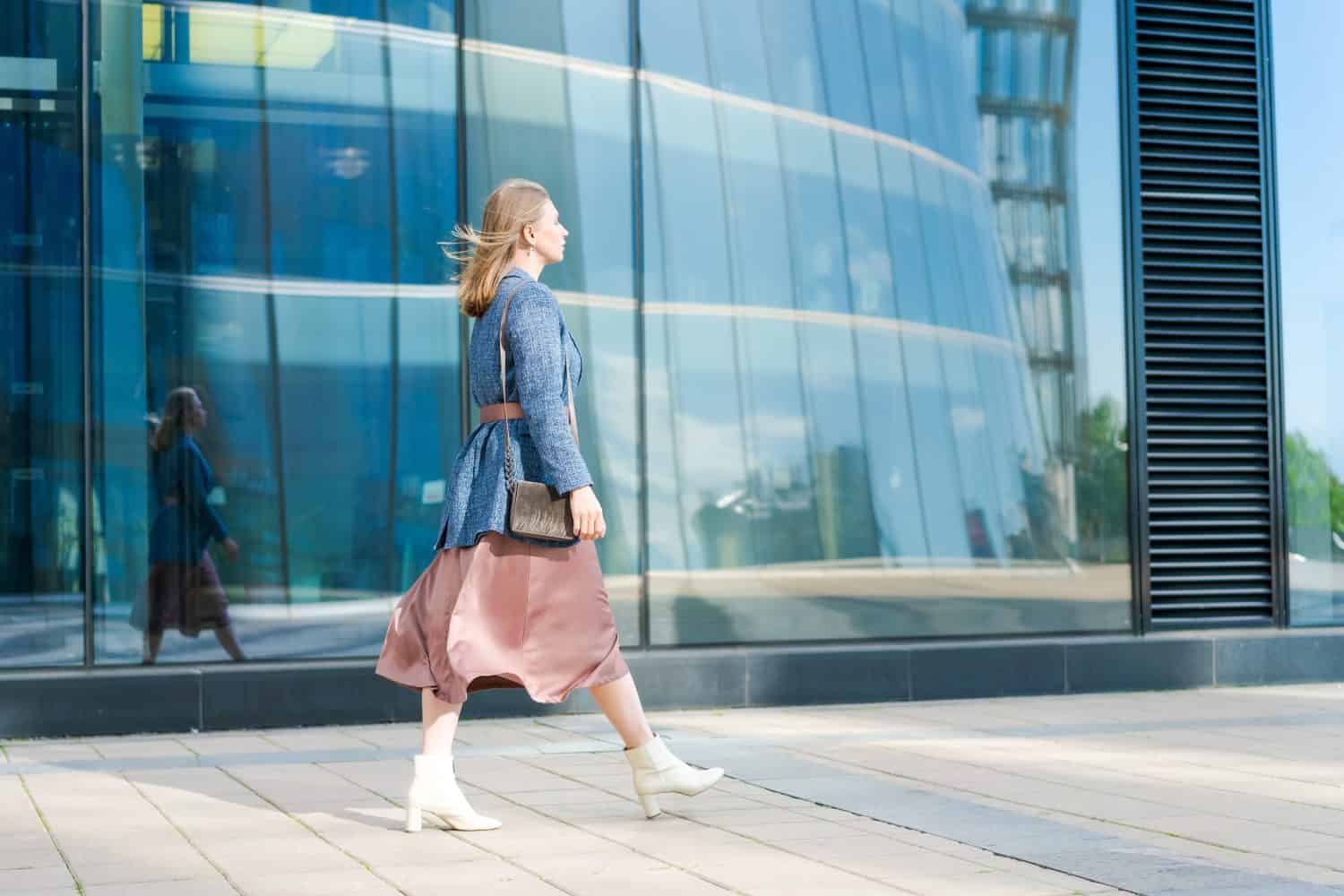 Successful happy woman on her way to work in street. Confident business woman with a smile in stylish clothes. Smiling lady steps on the background of the business center walk along the city street.