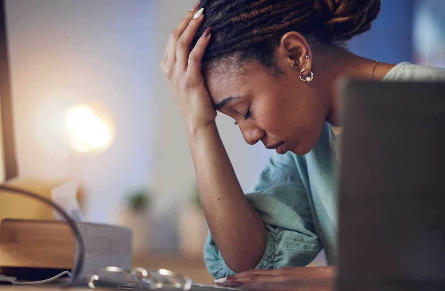 Business woman, depression and stress in an office at night working late on deadline. Tired African entrepreneur person with hands on head for pain, burnout or regret for mistake or fail at work