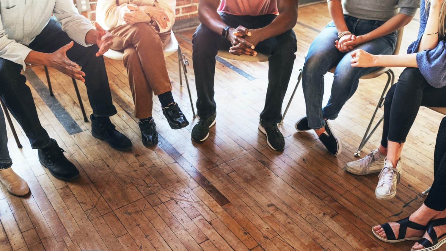 Diverse group sitting in a circle, engaged in discussion, wooden floor. Focus on hands and legs, showing interaction and communication. Diverse people sitting in a circle of support from community.