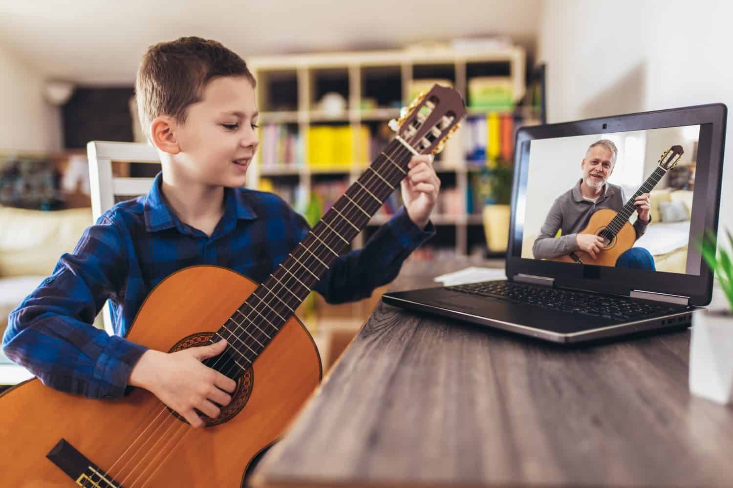 Focused boy playing acoustic guitar and watching online course on laptop while practicing at home. Online training, online classes.