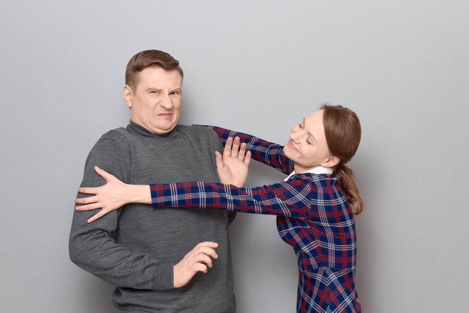 Studio shot of funny couple, happy cheerful woman is trying to hug man resisting and grimacing from displeasure, both are standing over gray background. Relationship concept, human emotions