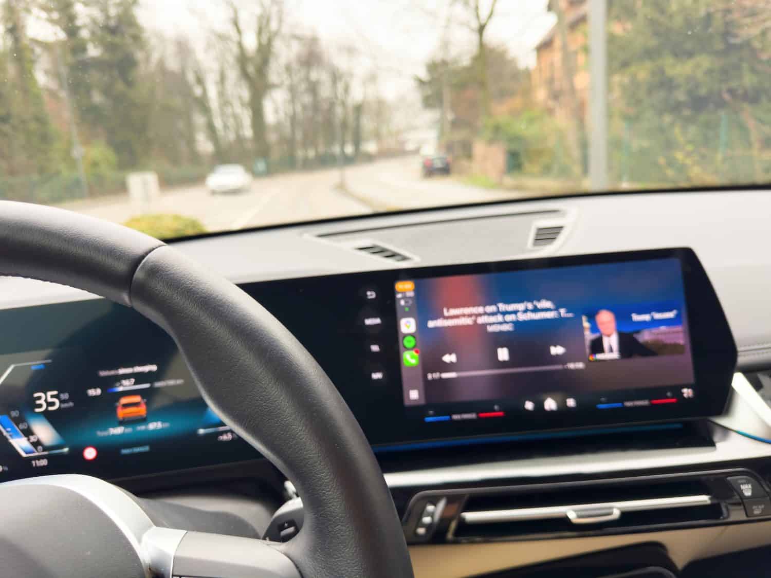Inside an electric car showing a large central display broadcasting defocused news content while the steering wheel and the driving screen are visible and the road outside is blurred.-
