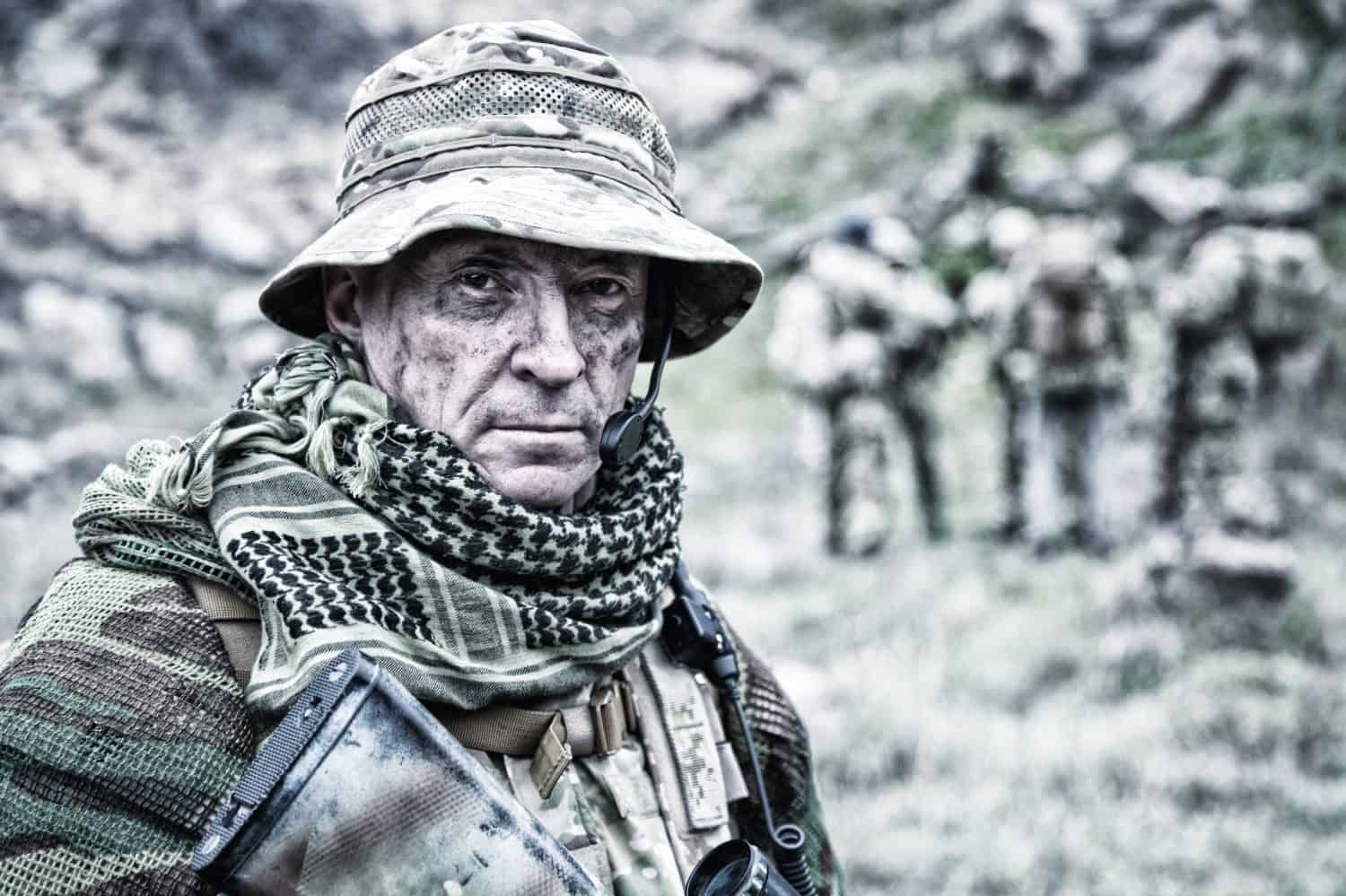 Close-up portrait of brutal commando veteran, experienced army commander or officer with dirty face, wearing camouflage bonnie, shemagh, tactical radio headset with microphone, looking in camera