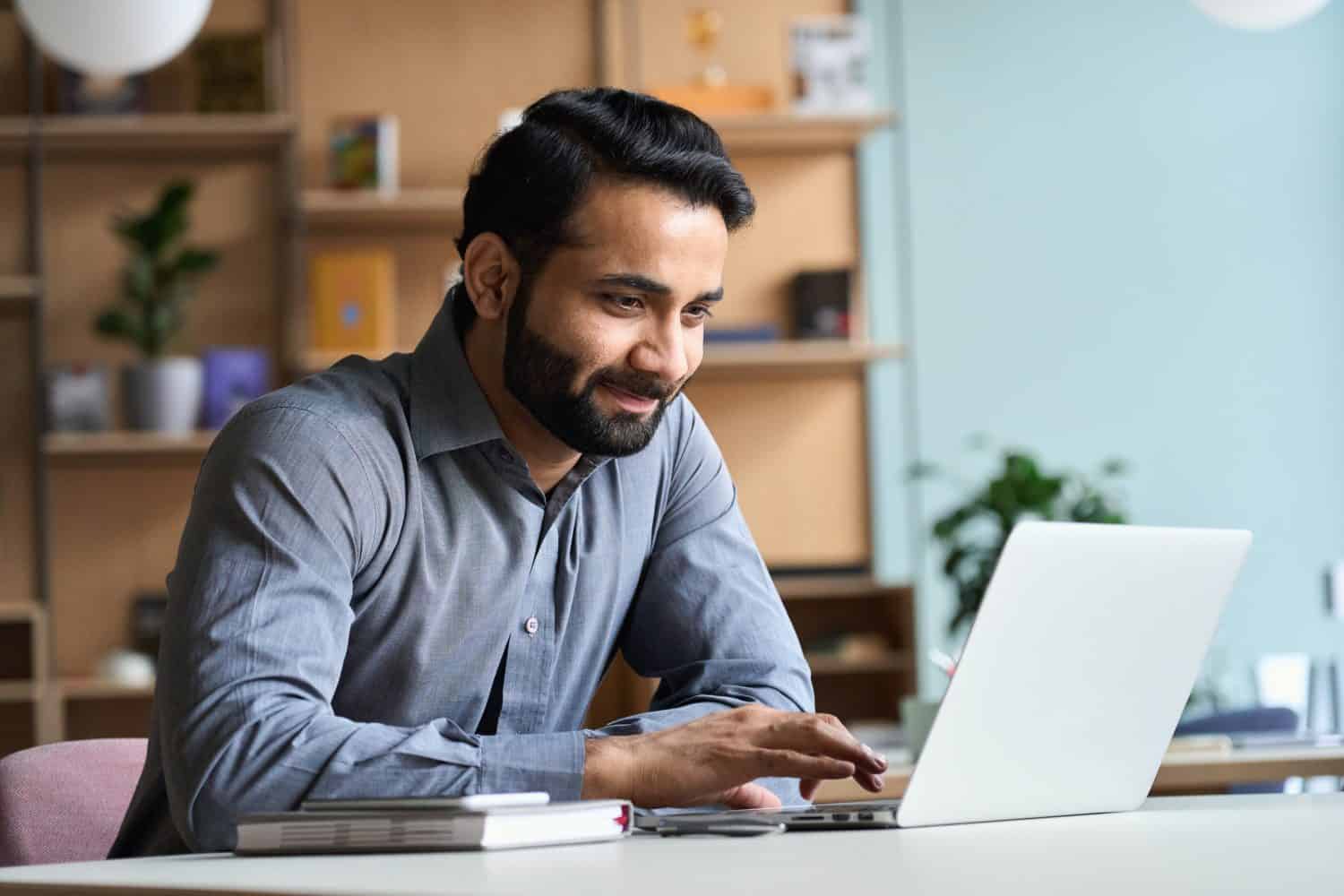 Smiling indian business man working on laptop at home office. Young indian student or remote teacher using computer remote studying, virtual training, watching online education webinar at home office.