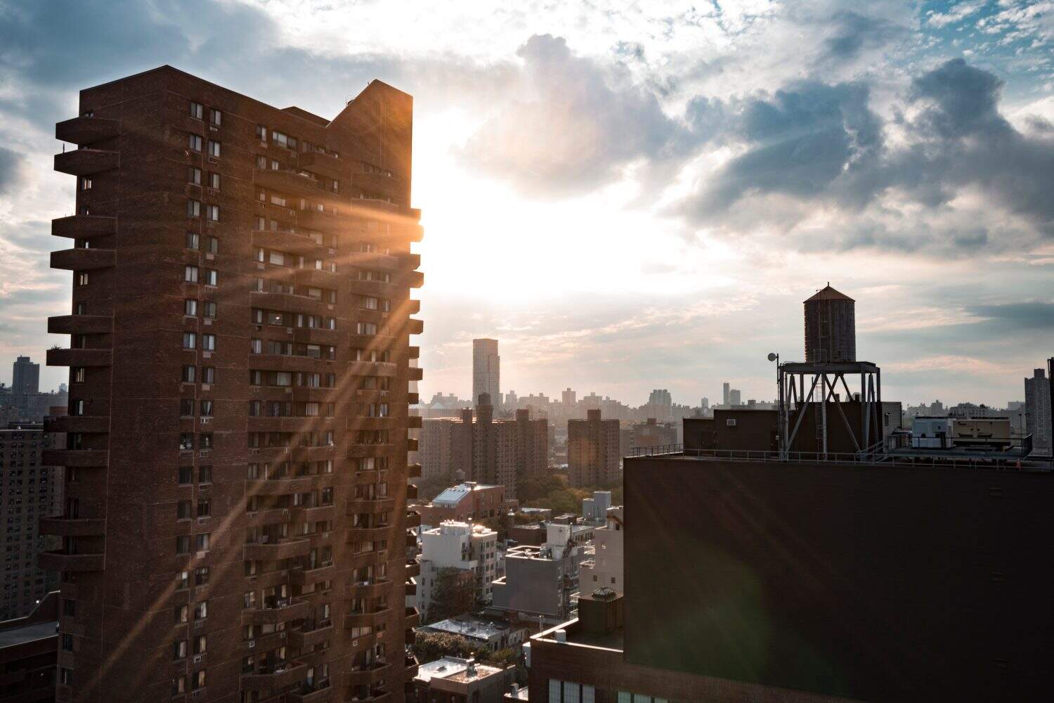 View from the 23rd floor of a apartment in east harlem, a water tank on the top of a building on the right and a big skyscraper on the left side. Between of this the sun comes through a cloudy sky.