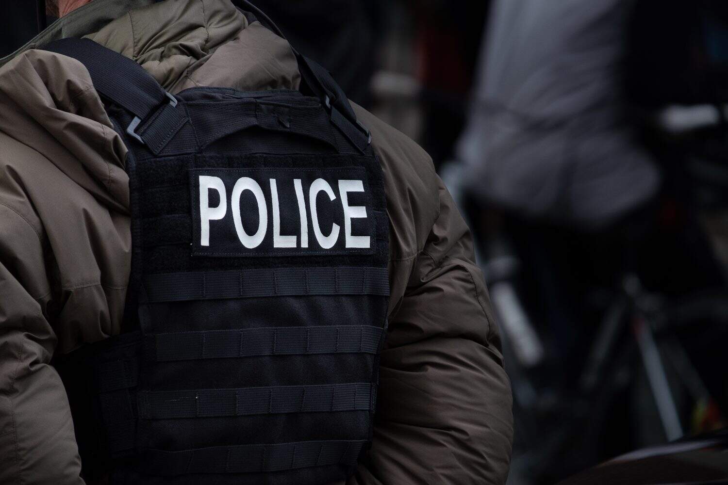 A male police officer stands in winter down-filled uniform clothing with a black bulletproof vest. The word police spelled out on the back of the vest. The officer has his hands folded behind his back