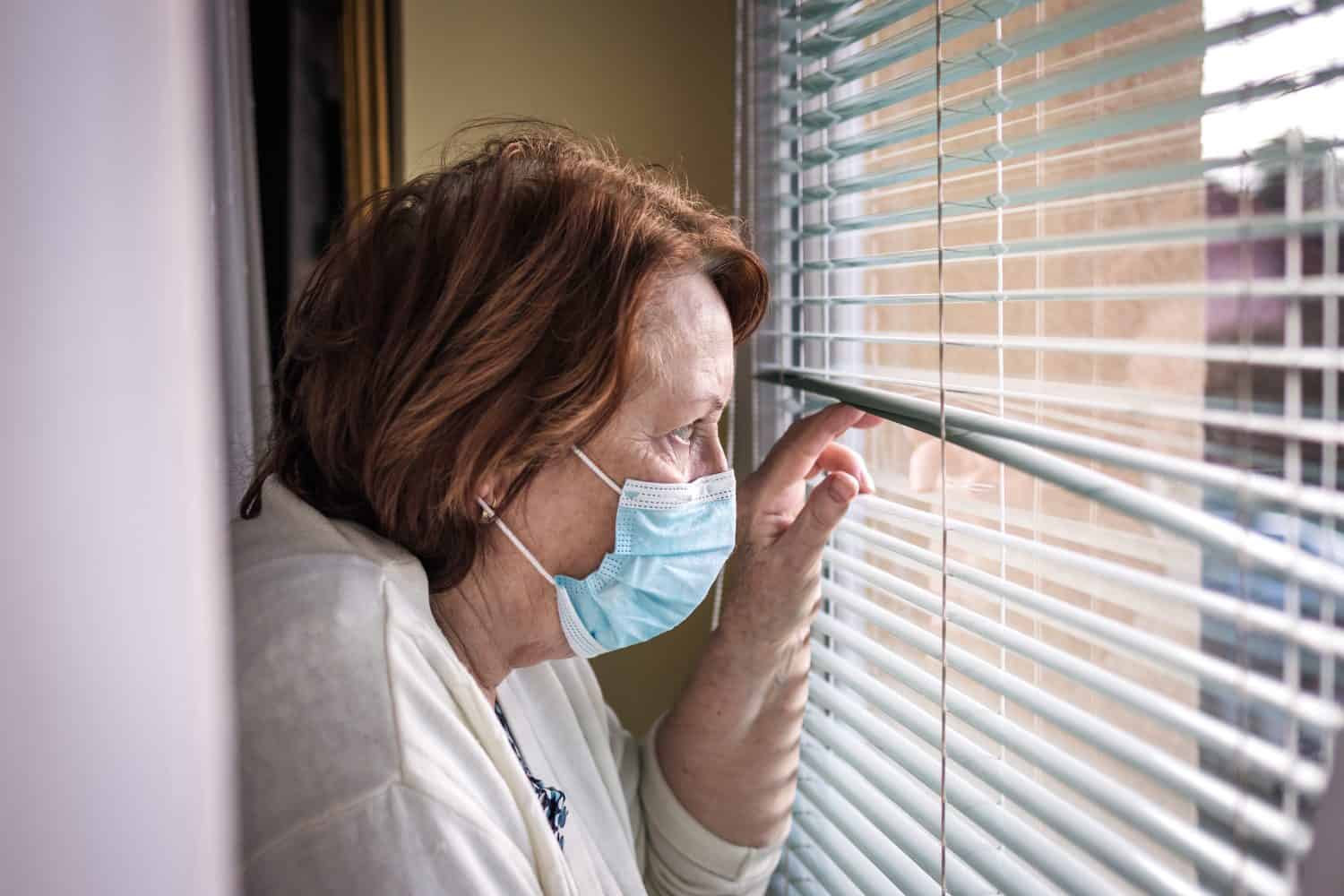 Lonely sad senior woman with face mask looking through window during covid-19 pandemic quarantine and lockdown