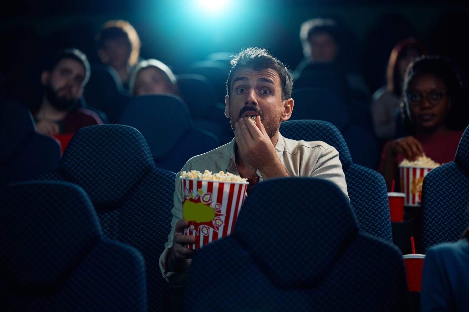 Young man eating popcorn while watching movie in disbelief in cinema.