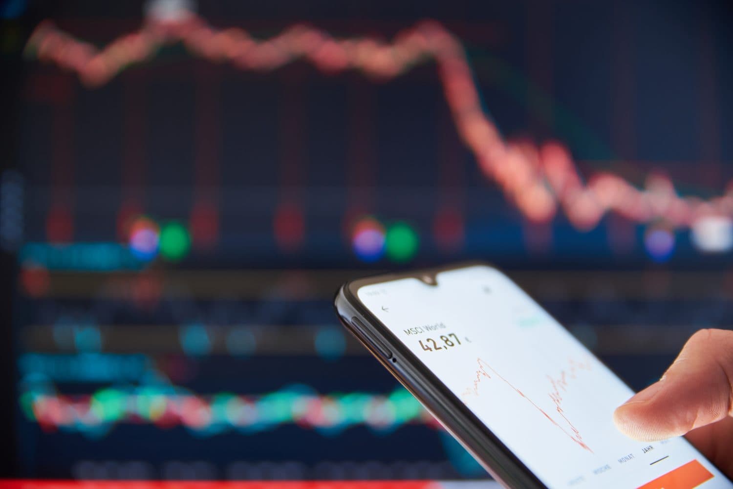 A person's hand holds a black smartphone displaying a financial charting application. The screen shows 'MSCI World 42,87' with a red line indicating a downward trend. In the blurred background, a large digital display also features multiple red, downward-sloping lines against a dark background, suggesting a significant market downturn.