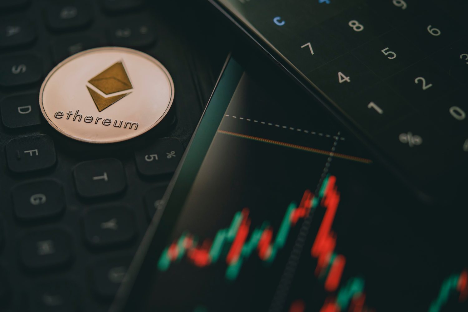 Close up of an Ethereum cryptocurrency coin near a monitor with candle stick graph chart and digital calculator.
