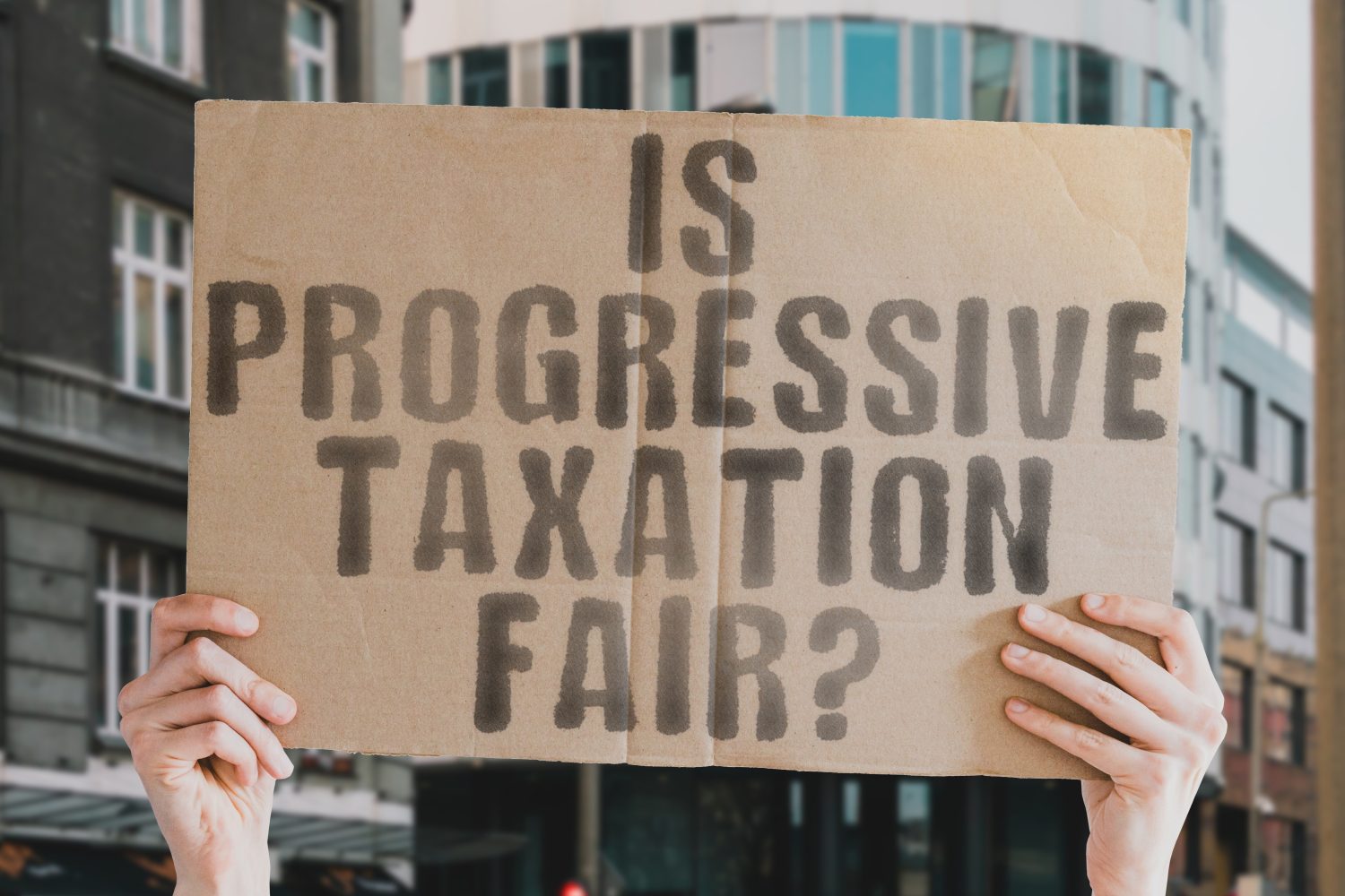 The question " Is progressive taxation fair? " is on a banner in men's hands with blurred background. Federal. Increase. Growing. Grow. Potential. Up. System. Trend. Return. Revenue. Sale. Office