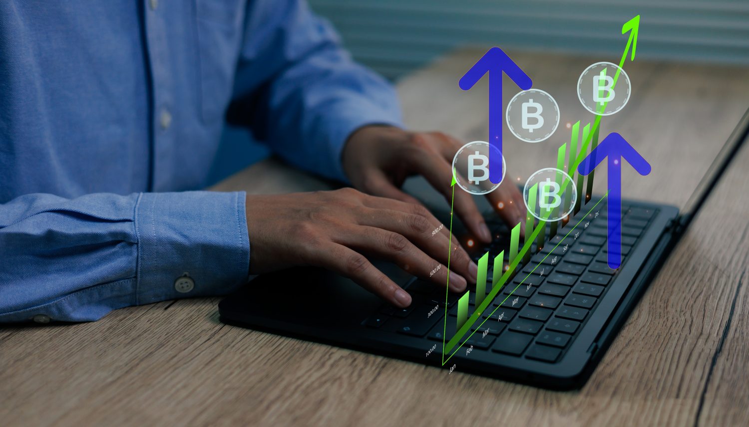 Close-up of a person's hands typing on a black laptop keyboard, with vibrant green and blue holographic digital overlays. The overlays show a rising bar chart, upward-pointing arrows, and several Bitcoin 'B' symbols, indicating financial growth and cryptocurrency market activity, against a wooden desk background.